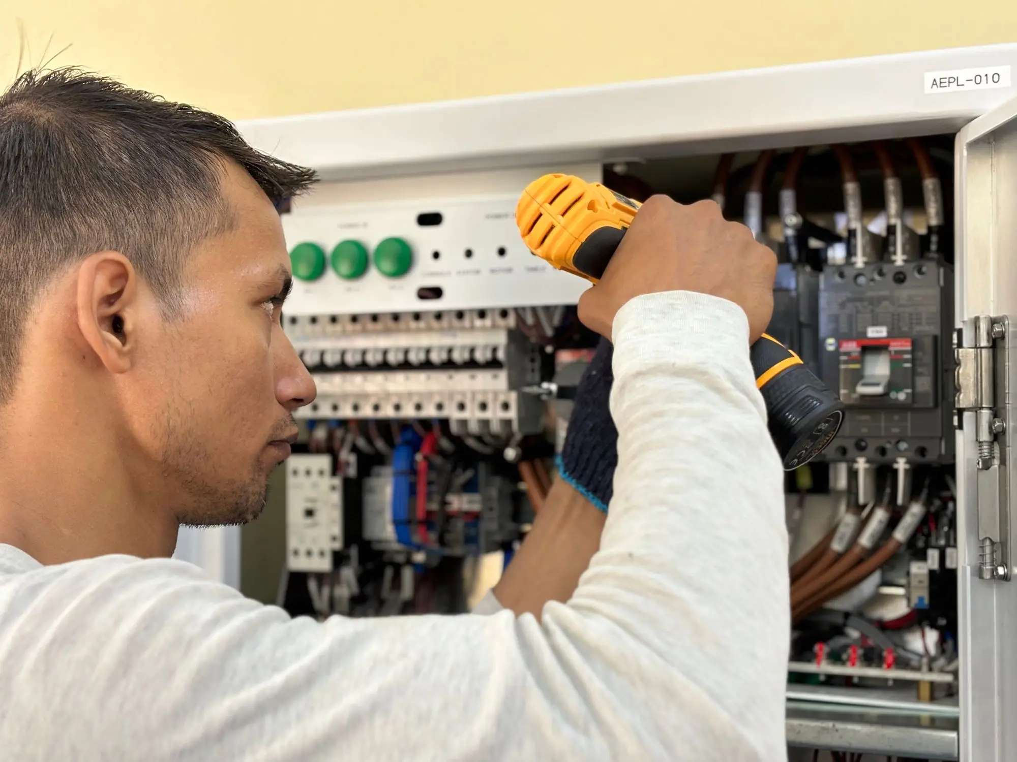 An electrician working at an open residential breaker panel with a cordless drill