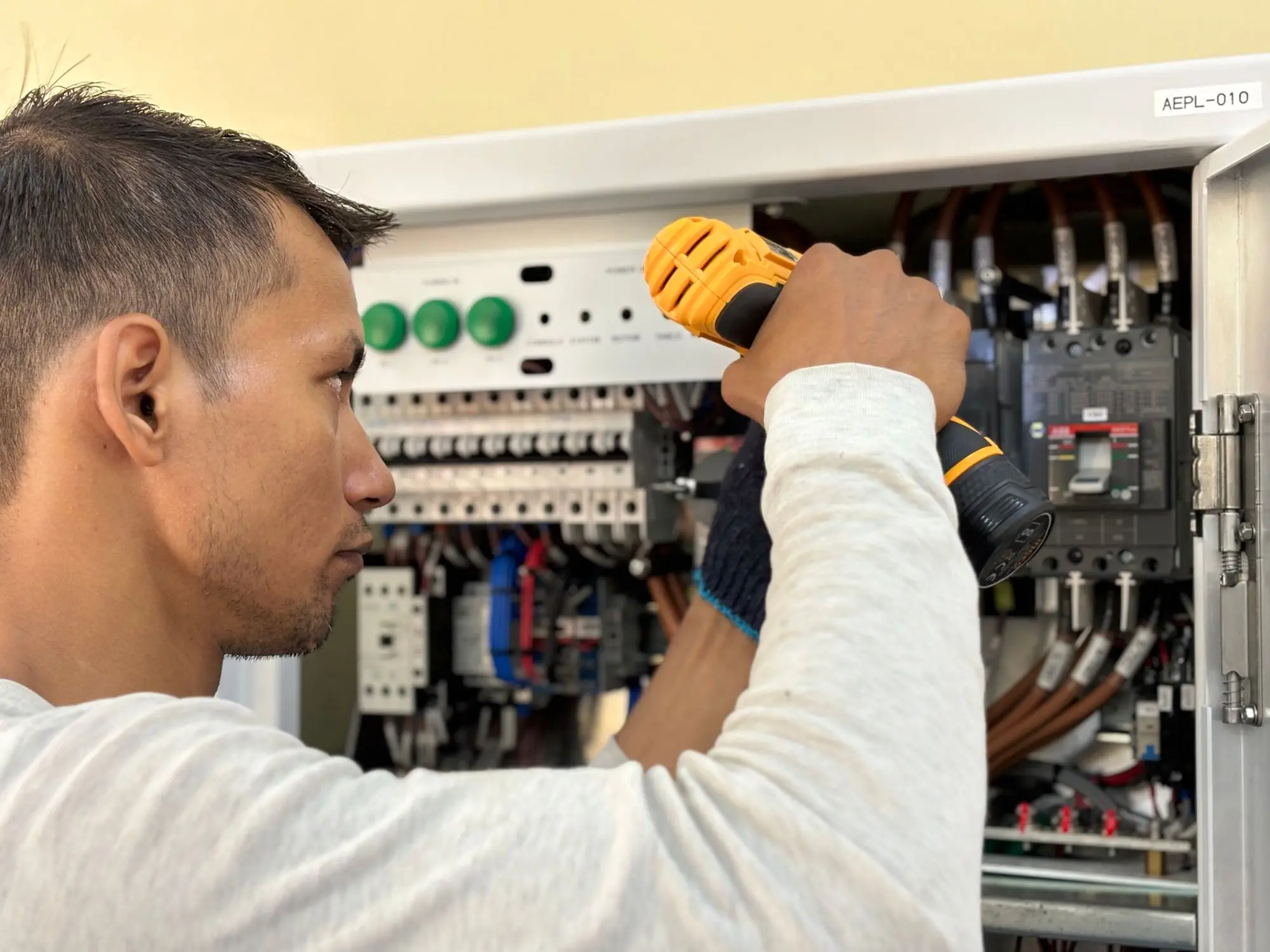 An electrician working at an open residential breaker panel with a cordless drill