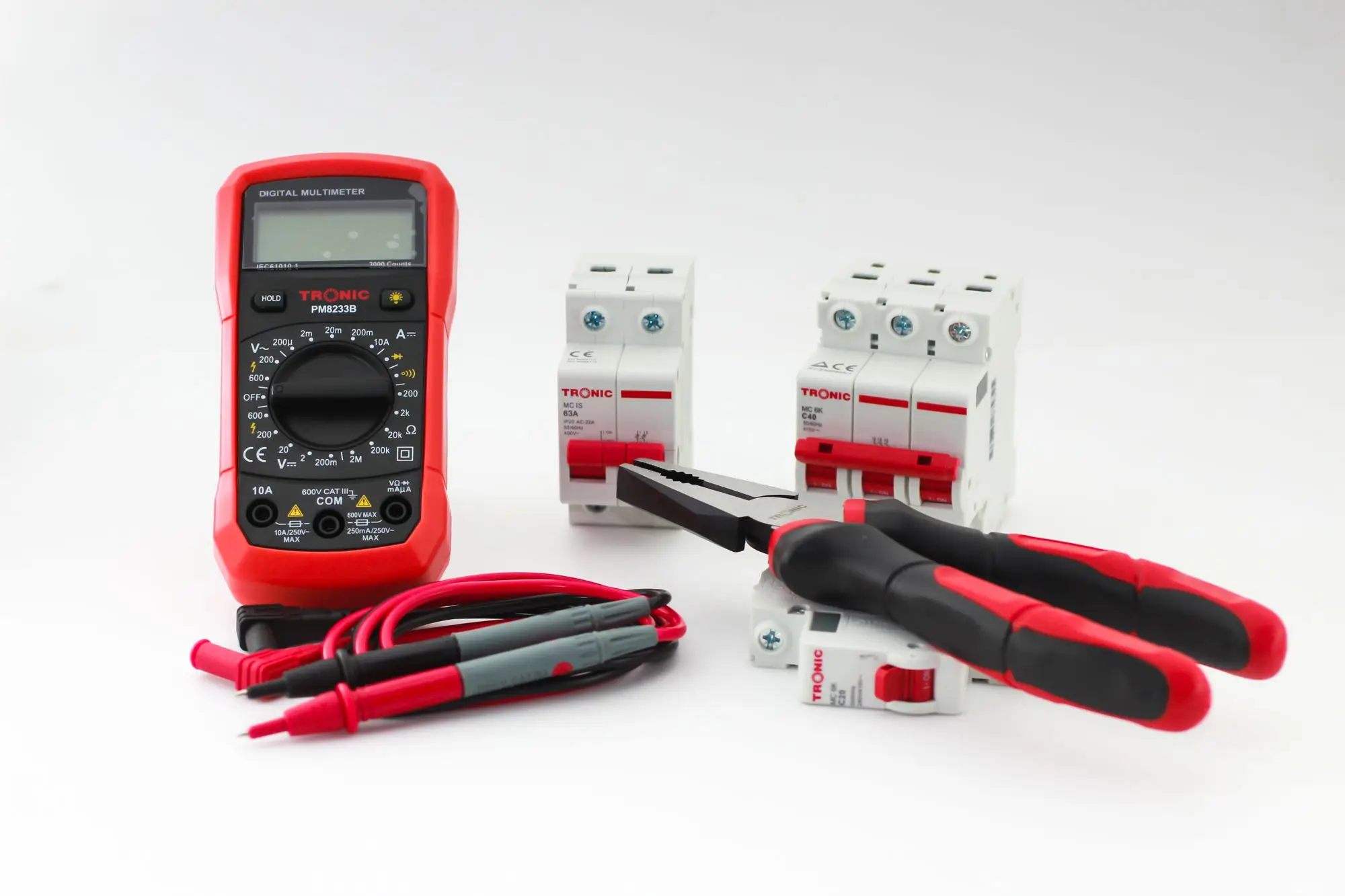 A digital multimeter lying beside hand tools and two circuit breakers on a white surface